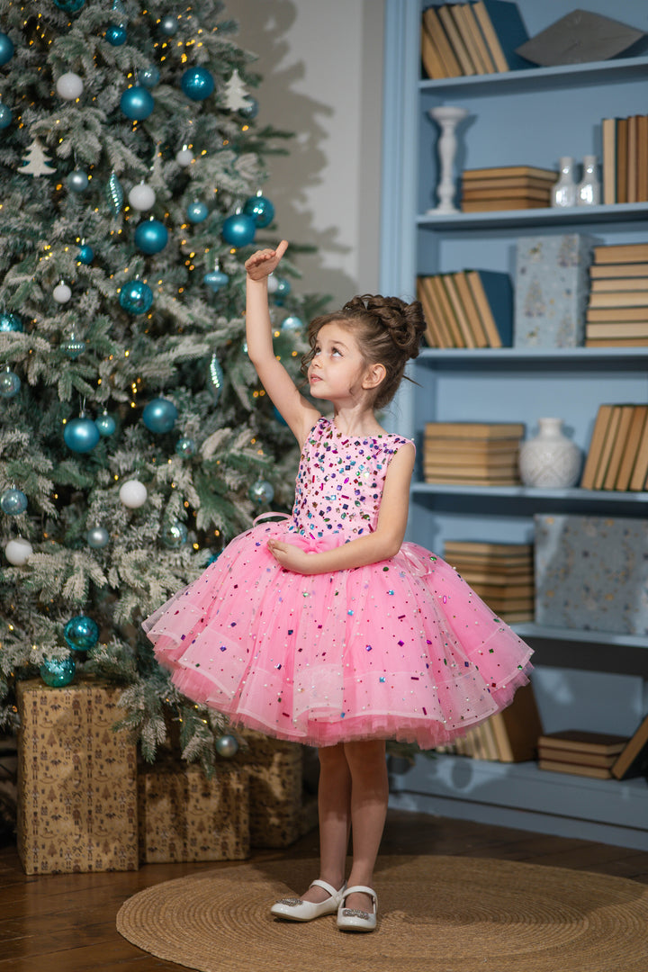 Baby girl in a pink dress standing next to a decorated Christmas tree.