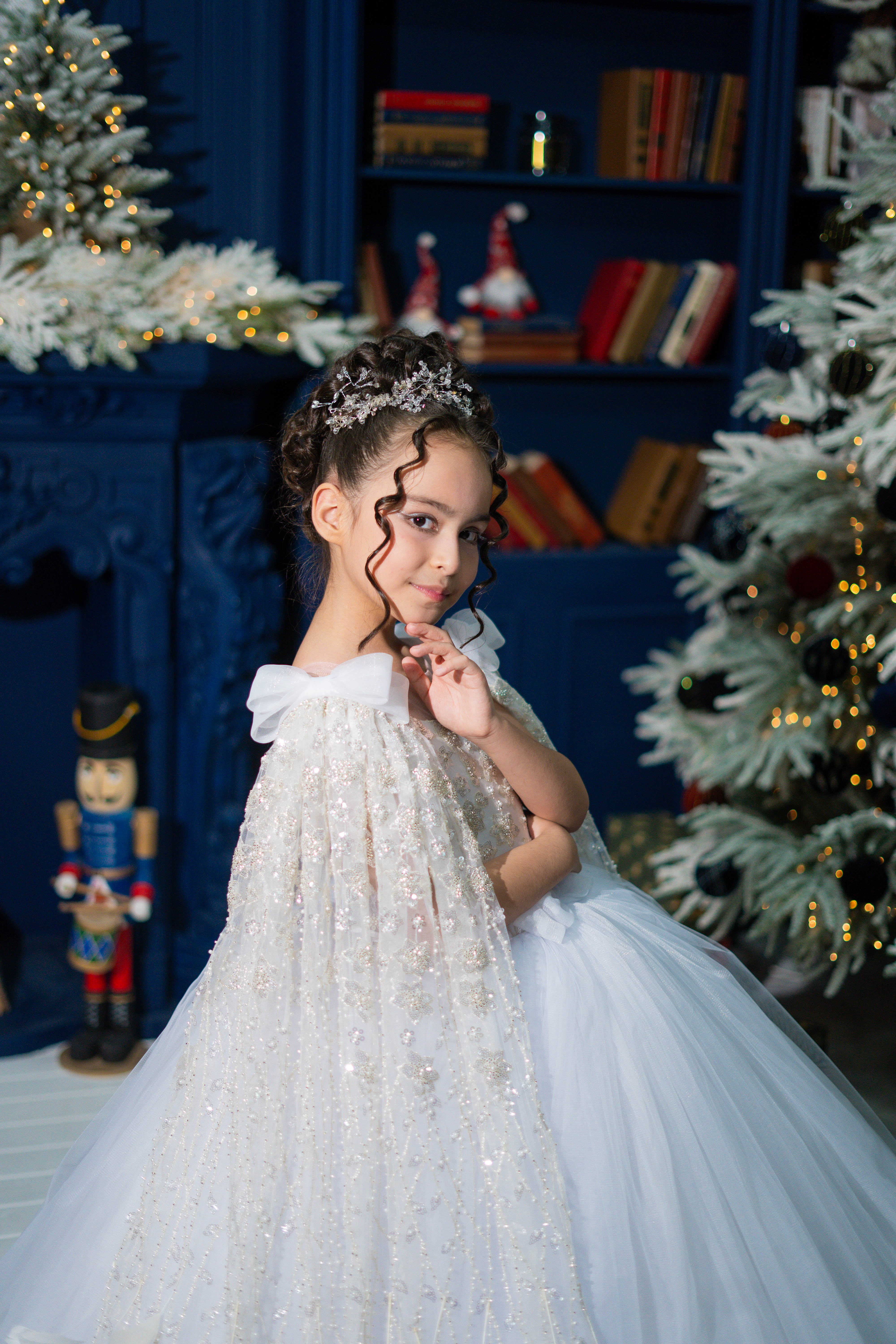 Young girl in a white dress with a bow standing in front of a decorated Christmas tree and bookshelf.