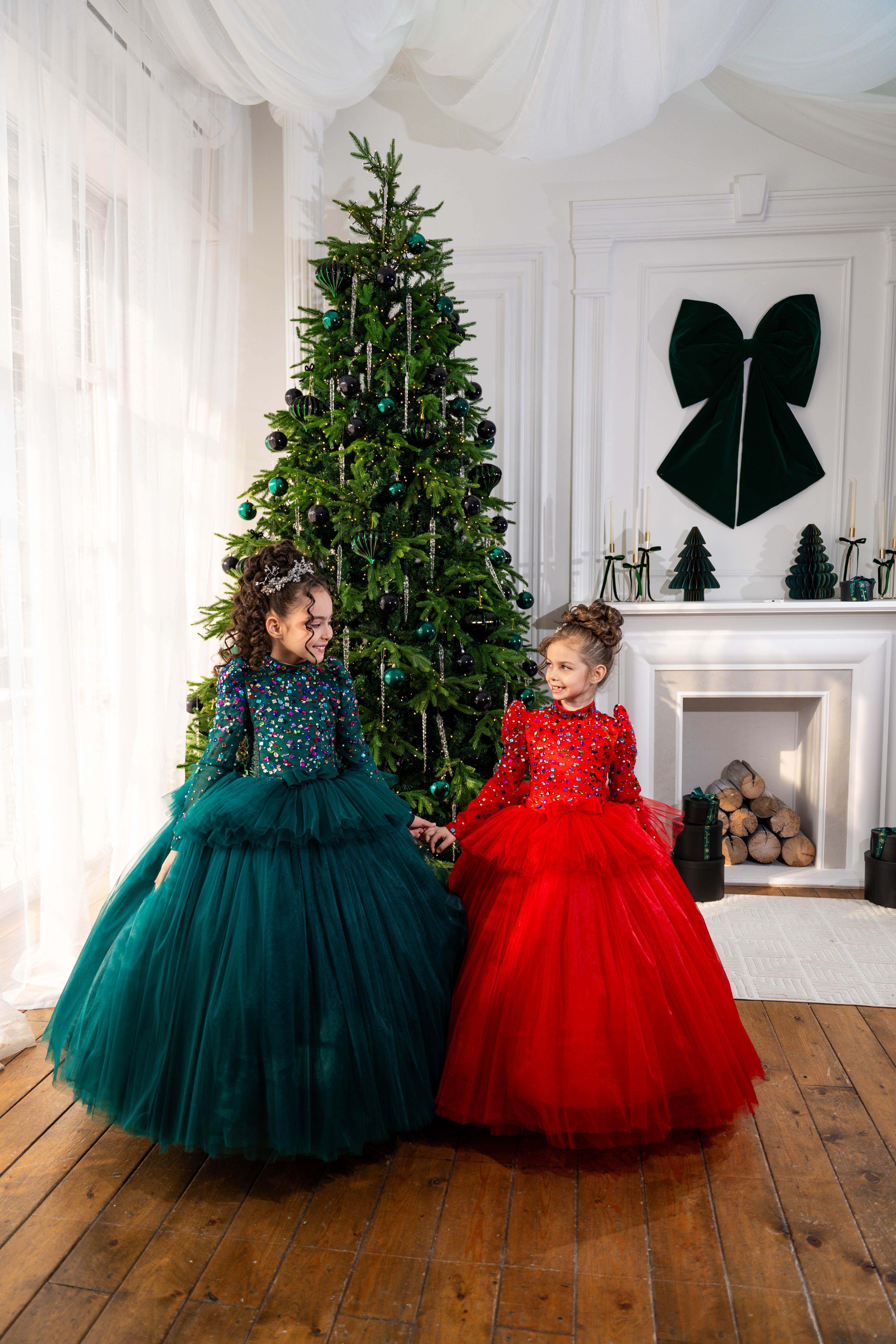 Two children in festive dresses sitting in front of a decorated Christmas tree in a room with a fireplace.