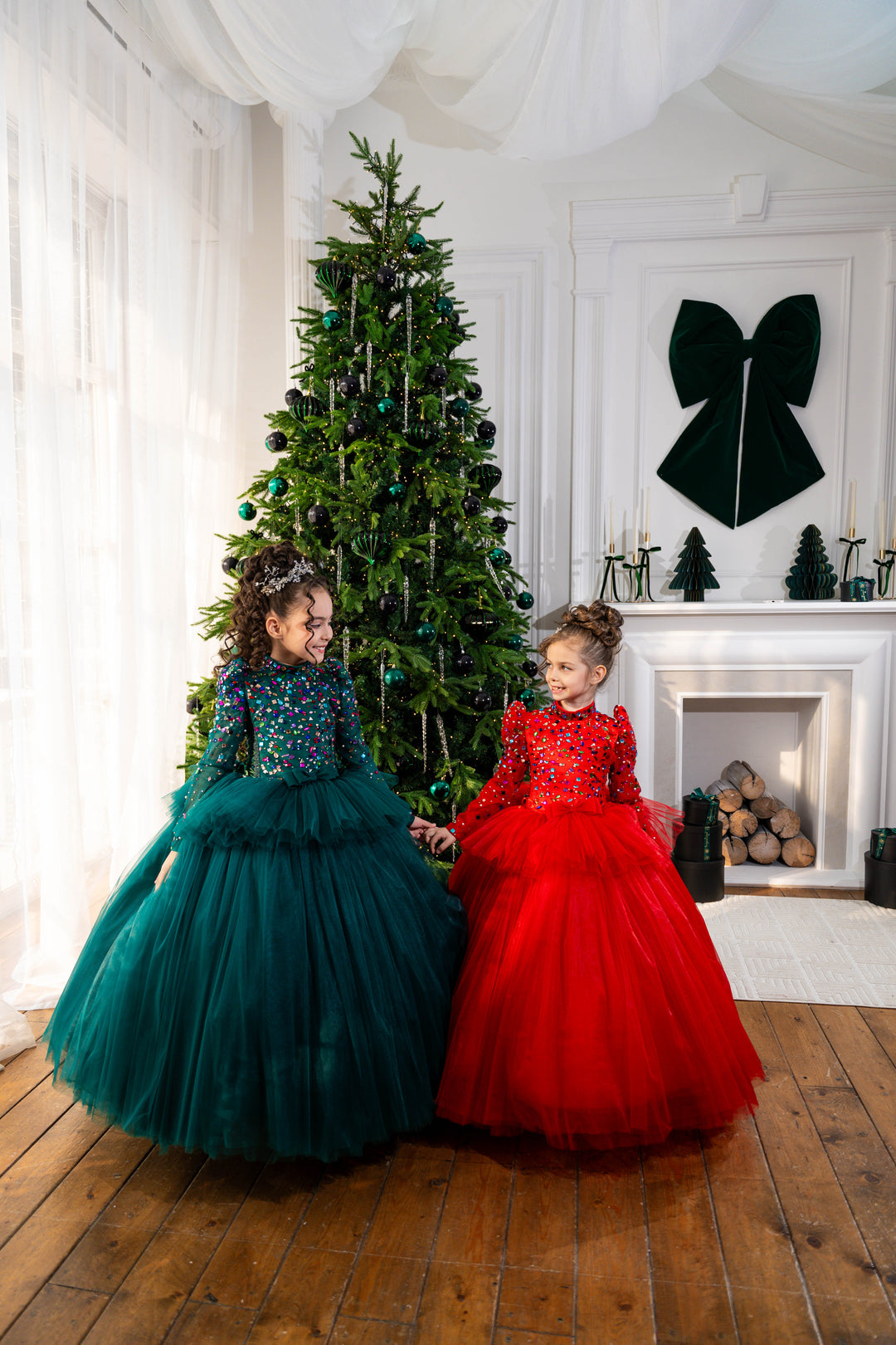 Two children in festive dresses sitting in front of a decorated Christmas tree in a room with a fireplace.