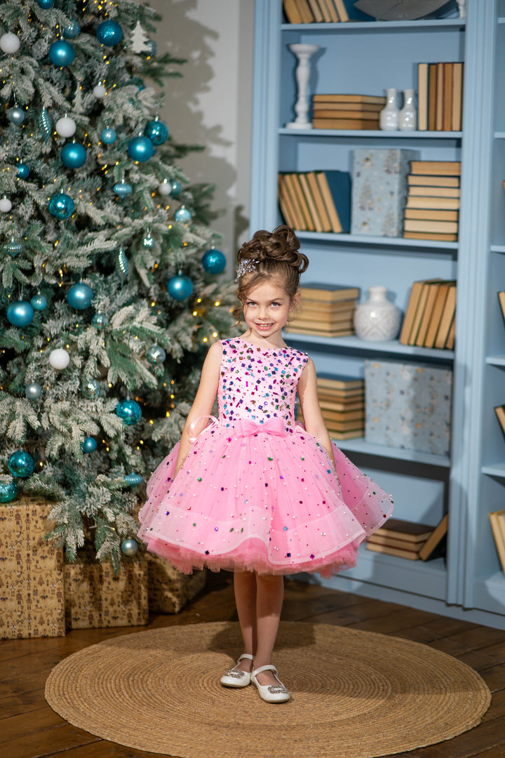 Baby girl in a pink dress standing in front of a decorated Christmas tree and bookshelf.