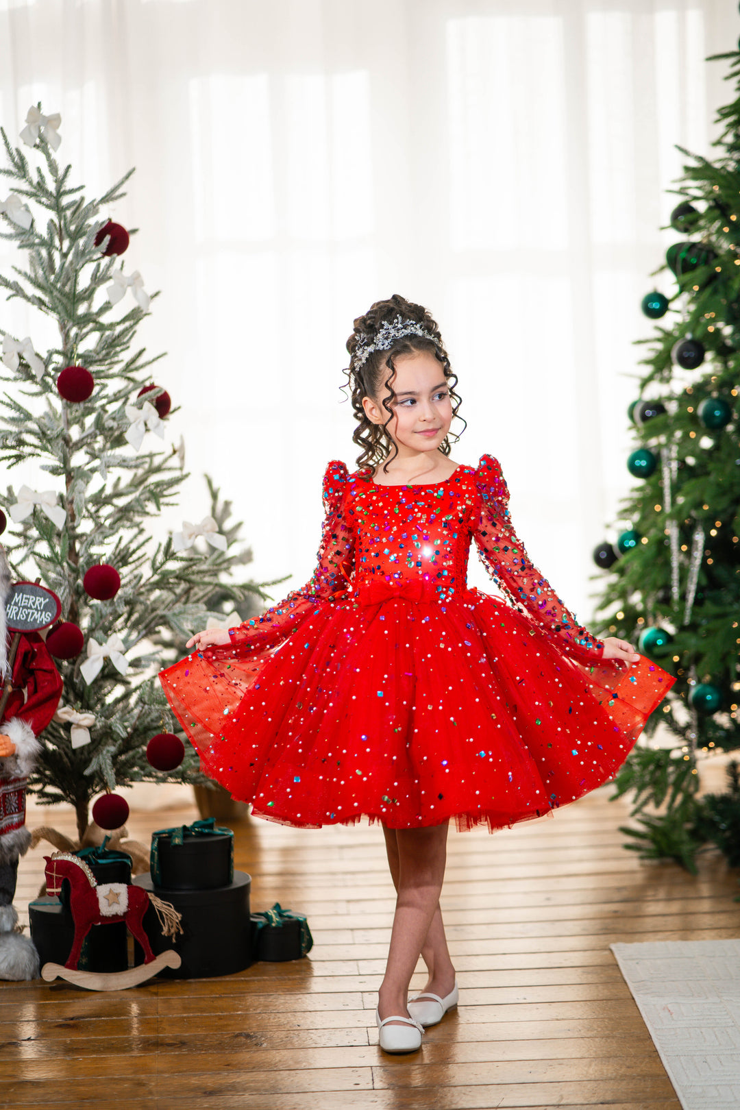 Baby girl in a red dress standing between two decorated Christmas trees.
