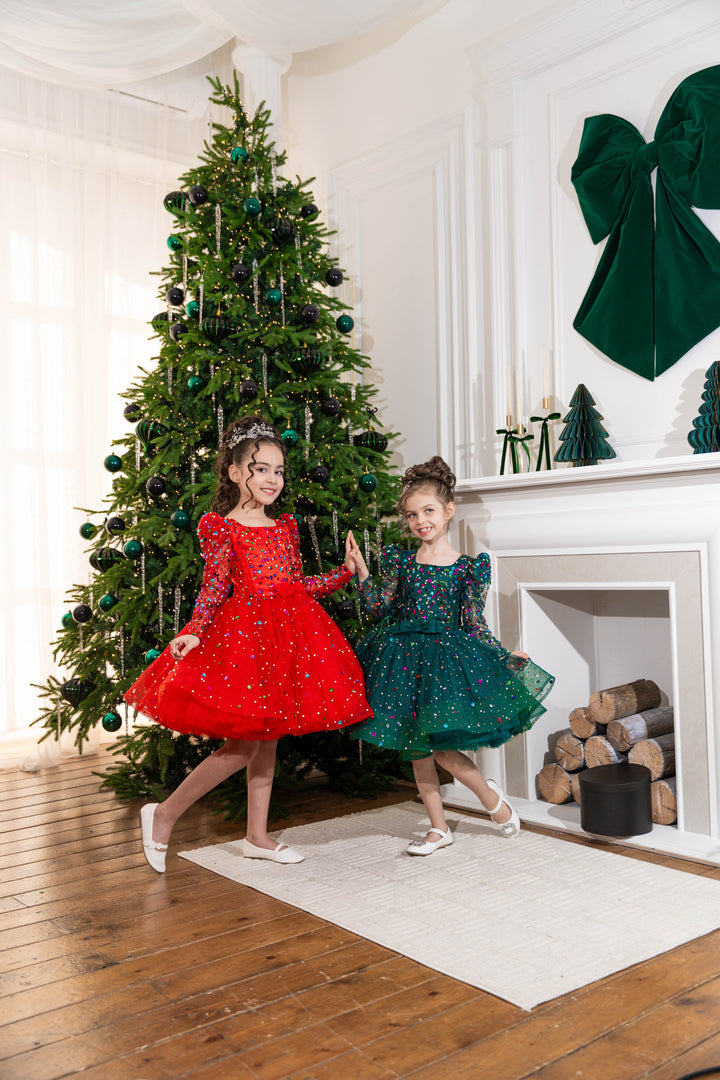 Two children in red and green dresses standing in front of a decorated Christmas tree.