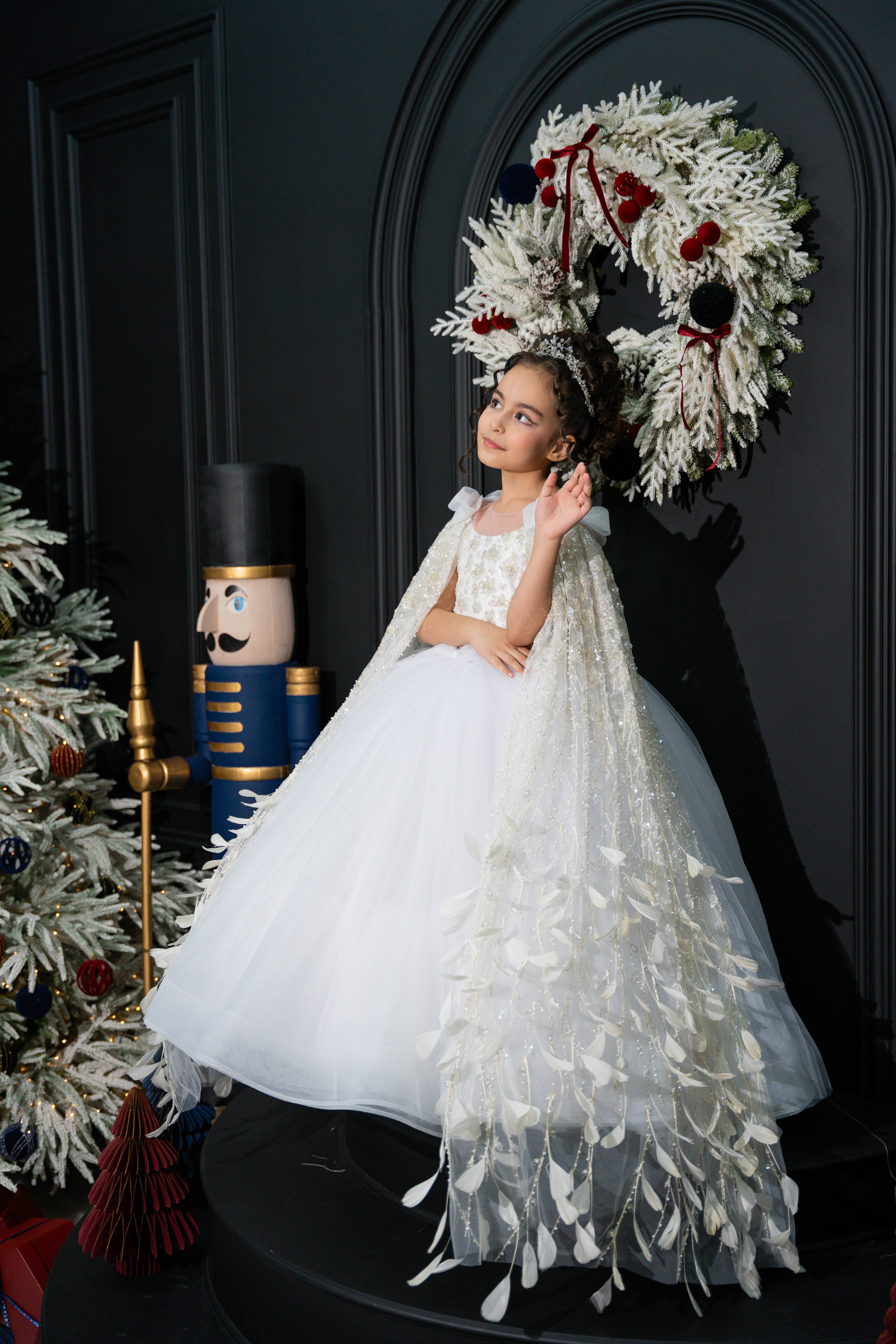 A child in a white dress with a decorative wreath and Christmas tree in the background