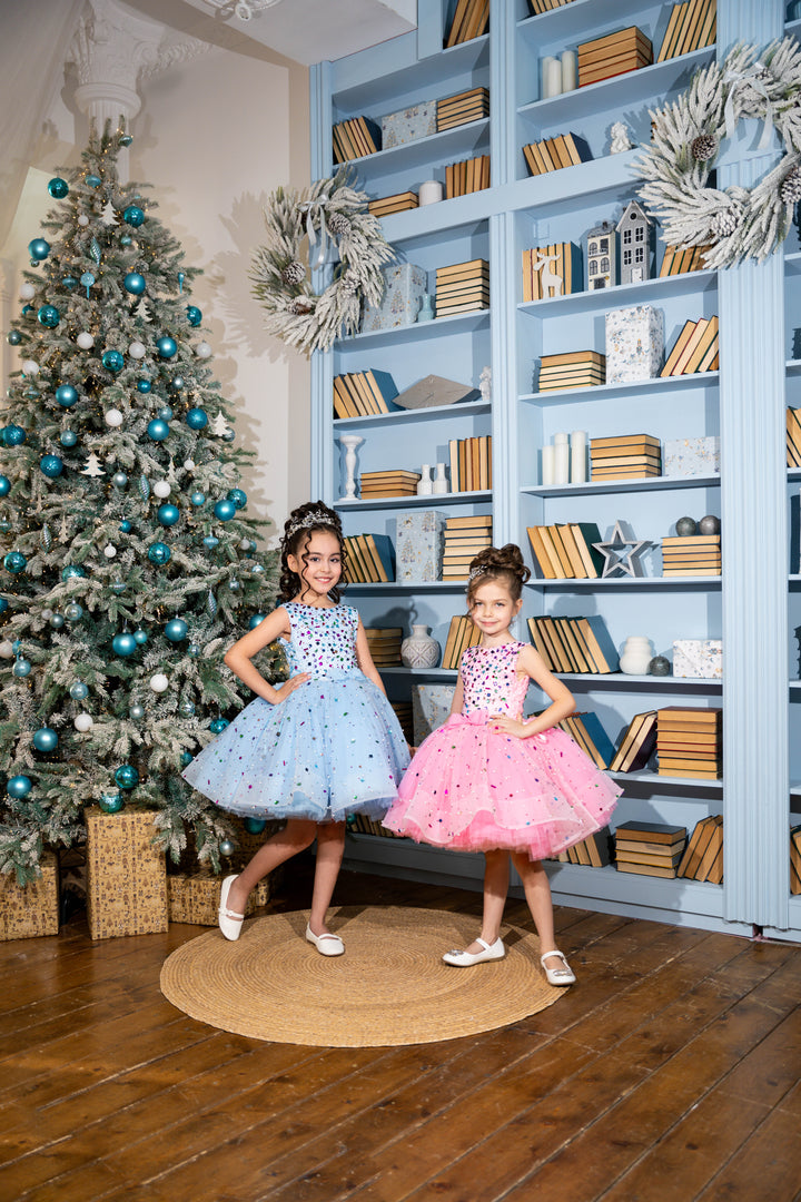 Two young girls in dresses standing in a room with a decorated Christmas tree and bookshelf.