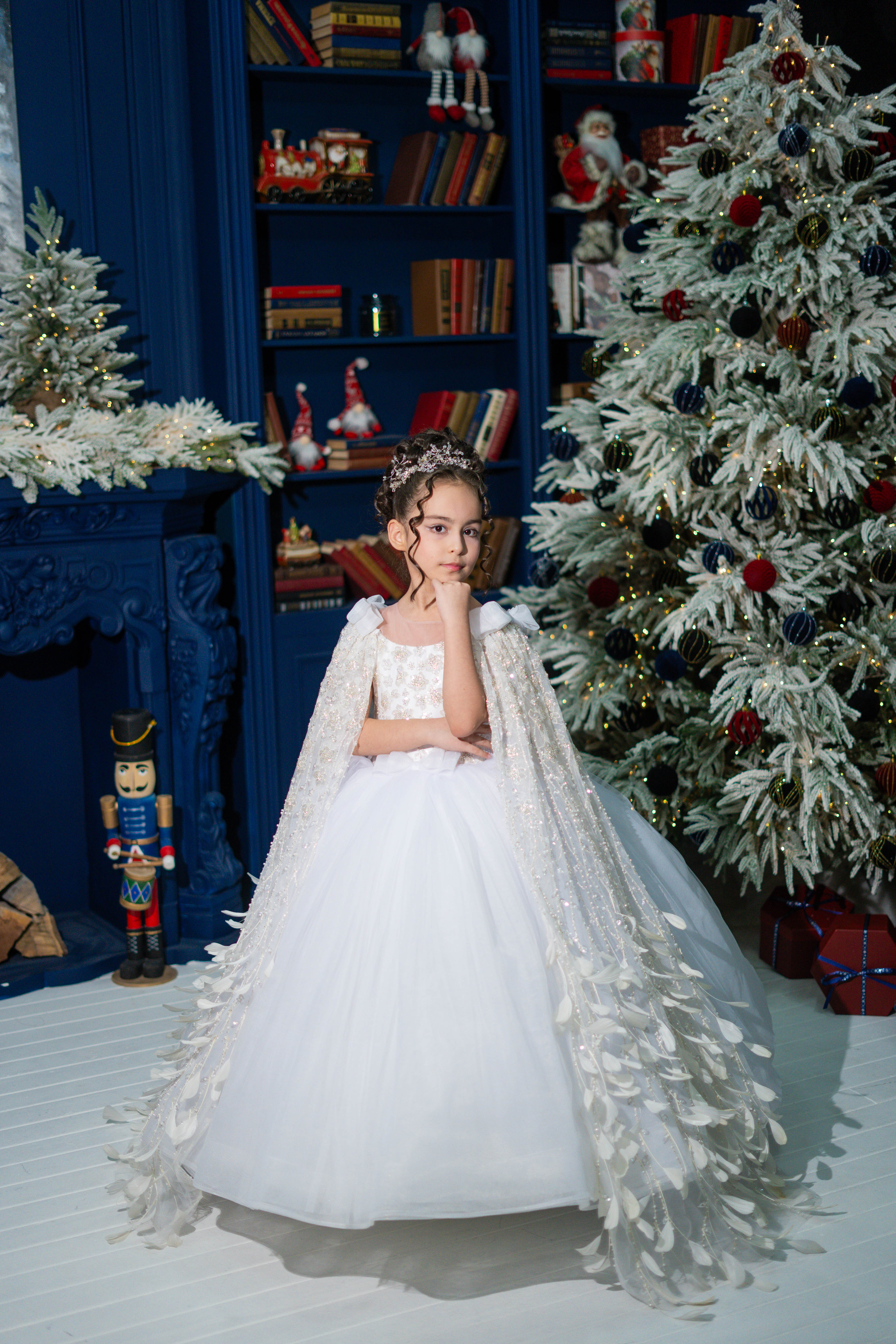 Child in a white dress with a floral headband standing in front of a decorated Christmas tree and bookshelf.