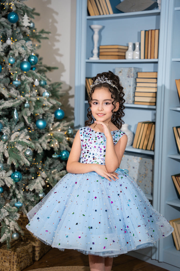Young girl in a light blue dress with colorful polka dots standing next to a decorated Christmas tree.