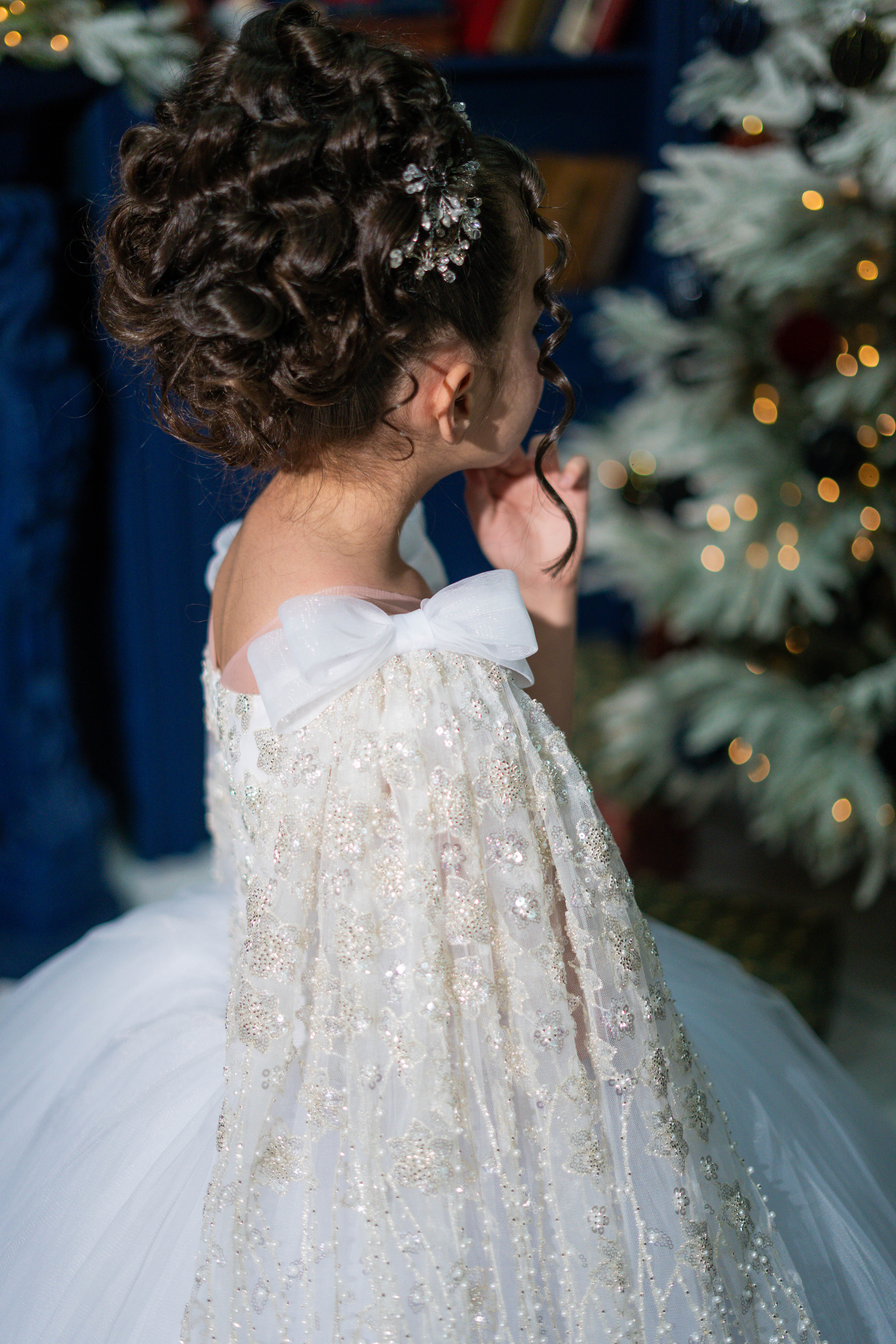 Child in a sparkling white dress with braided hair and floral accessory, Christmas tree in the background