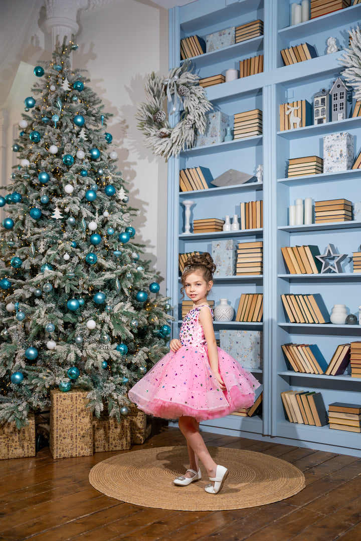 Baby girl in a pink dress standing in front of a decorated Christmas tree and bookshelves.